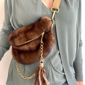 May include: A brown fur fanny pack with a gold chain strap and a brown fur tassel. The bag is worn over a person's shoulder.