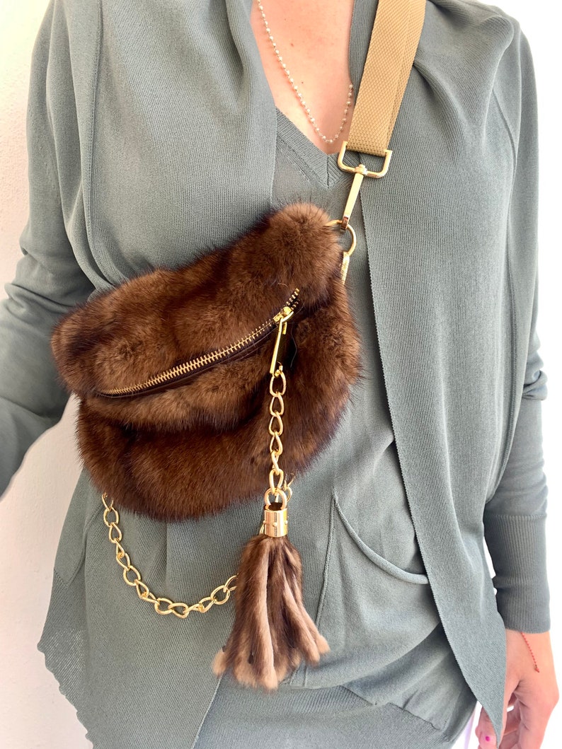 May include: A brown fur fanny pack with a gold chain strap and a brown fur tassel. The bag is worn over a person's shoulder.