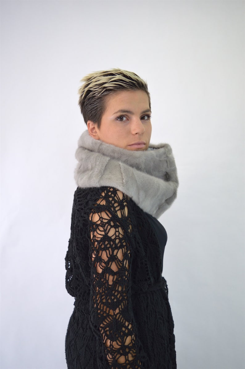 May include: A woman with short blonde hair is wearing a grey fur scarf and a black crochet top.