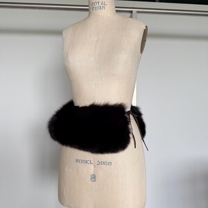 May include: A dark brown fur belt is wrapped around a vintage dress form. The belt is made of thick, plush fur and is tied with a thin, dark brown leather cord. The dress form is beige and has the words "ROYAL FORM" and "MODEL 2000" printed on it.