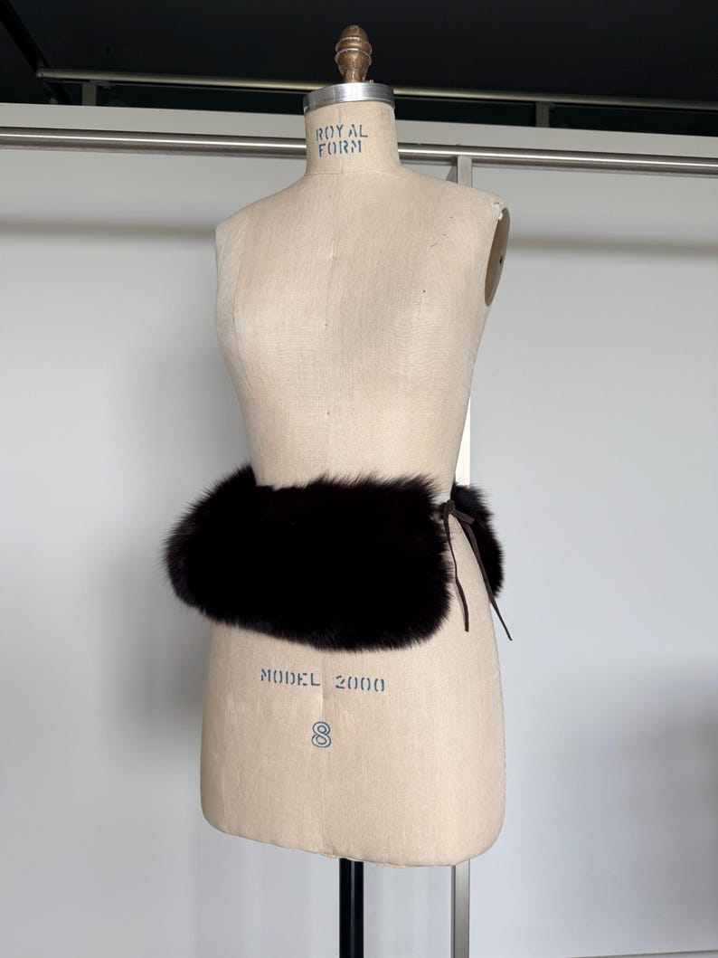 May include: A dark brown fur belt is wrapped around a vintage dress form. The belt is made of thick, plush fur and is tied with a thin, dark brown leather cord. The dress form is beige and has the words "ROYAL FORM" and "MODEL 2000" printed on it.