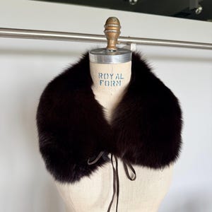 May include: A dark brown fur collar with a tie closure, displayed on a mannequin. The collar is thick and plush, with a rich, deep color. The mannequin has the text "ROYAL FORM" printed on it.