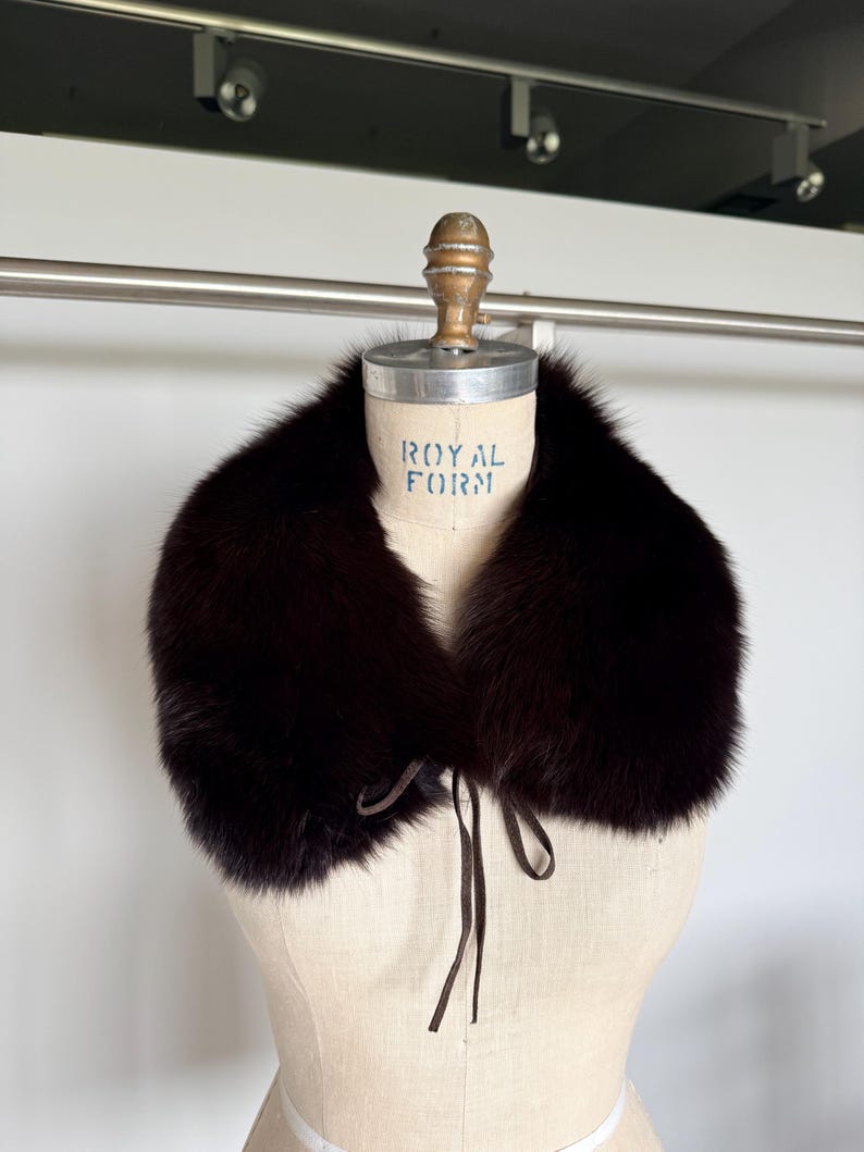 May include: A dark brown fur collar with a tie closure, displayed on a mannequin. The collar is thick and plush, with a rich, deep color. The mannequin has the text "ROYAL FORM" printed on it.
