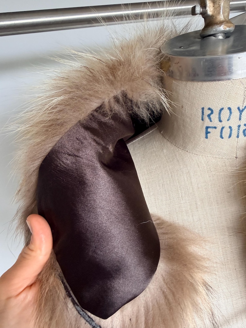 Op de afbeelding: Close-up van een kledingkraag met een luxe bontafwerking in beige en lichtbruine tinten. De binnenkant van de kraag is gevoerd met een gladde, donkerbruine stof. Het kledingstuk wordt op een mannequin getoond.
