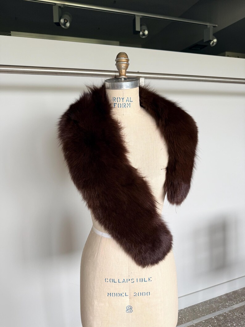May include: A dark brown fur stole draped over a dress form. The stole is a rich, deep brown color and appears soft and luxurious. The dress form is a light beige color, with text that reads "ROYAL FORM" and "COLLAPSIBLE MODEL 2000".