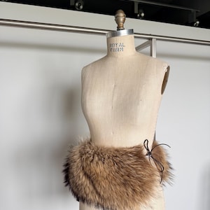 Op de afbeelding: Een bruine bontkraag met een donkerbruine striksluiting wordt getoond op een vintage paspop. De kraag is gemaakt van zacht, pluizig bont en is ontworpen om om de nek te worden gedragen.