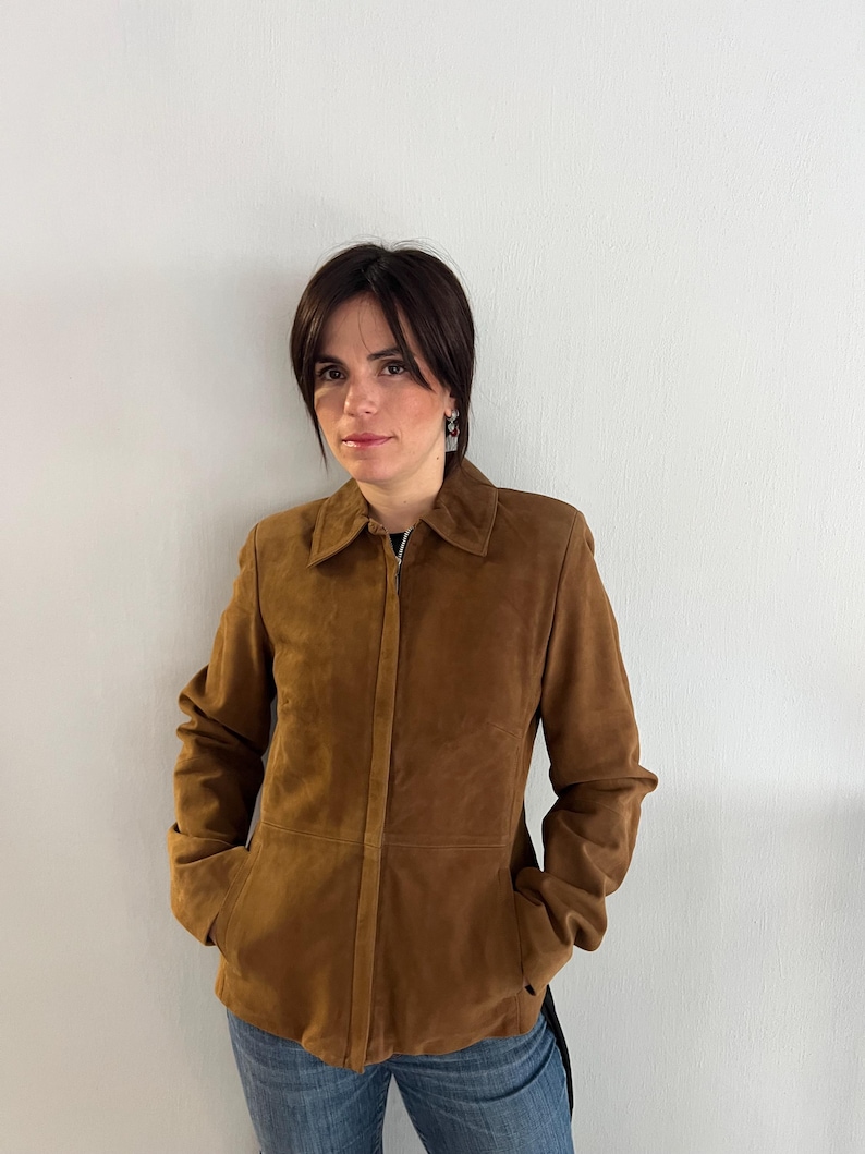 May include: A brown suede jacket with a collared neckline and a full-length zip. The jacket features two front pockets and is worn with blue jeans. The jacket's colour is a warm, earthy tone.