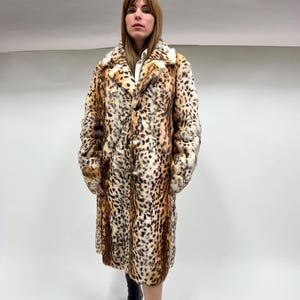 May include: A long, faux fur coat with a leopard print pattern. The coat features a wide collar and a button-down front. The colors include shades of brown, tan, and black. The coat is knee-length and worn with black boots.