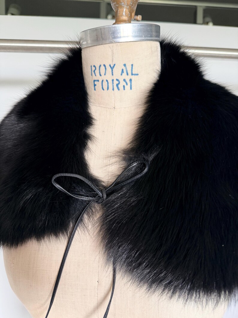 May include: A black fur collar with a leather tie closure. The collar is displayed on a mannequin with the text "ROYAL FORM" printed on it. The fur appears soft and luxurious.