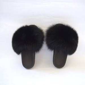 K&ouml;nnte beinhalten: Ein Paar schwarze Fellslipper. Die Slipper haben einen flauschigen schwarzen Pelzbesatz am oberen Rand des Slippes.