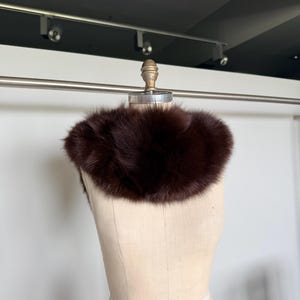 May include: A dark brown fur collar displayed on a mannequin. The collar is a rich, deep shade of brown, with a fluffy, textured appearance. The mannequin is a neutral beige color, and the background is a simple white wall.