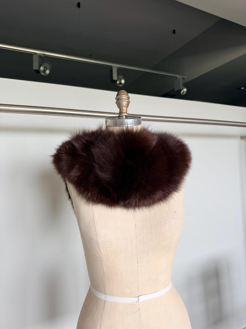 May include: A dark brown fur collar displayed on a mannequin. The collar is a rich, deep shade of brown, with a fluffy, textured appearance. The mannequin is a neutral beige color, and the background is a simple white wall.