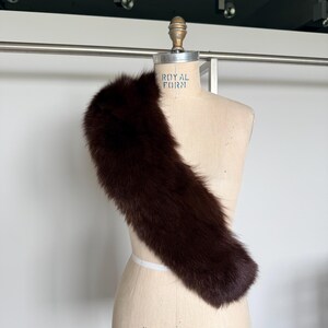 May include: A dark brown fur stole draped over a vintage dress form. The stole is long and fluffy, with a rich, deep color. The dress form is a light beige color, with the words "ROYAL FORM" and "COLLAPSIBLE MODEL 2000" printed on it.