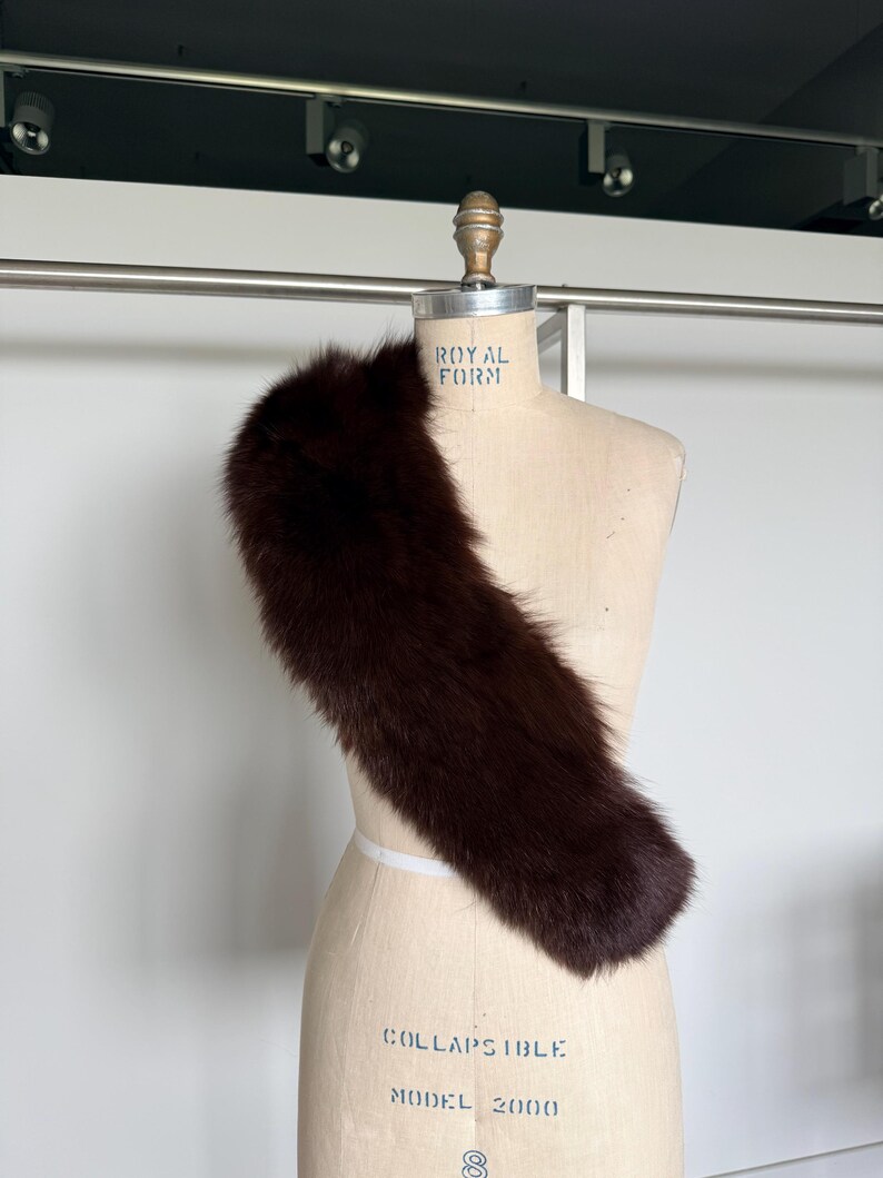 May include: A dark brown fur stole draped over a vintage dress form. The stole is long and fluffy, with a rich, deep color. The dress form is a light beige color, with the words "ROYAL FORM" and "COLLAPSIBLE MODEL 2000" printed on it.