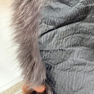 以下が含まれることがあります： テクスチャ加工された表面を持つ濃いグレーの衣服のクローズアップ。ふわふわのライトグレーの毛皮でトリミングされています。毛皮は厚くて柔らかく、生地のしわくちゃの外観とは対照的です。この衣服はアウターウェアの一部のようです。