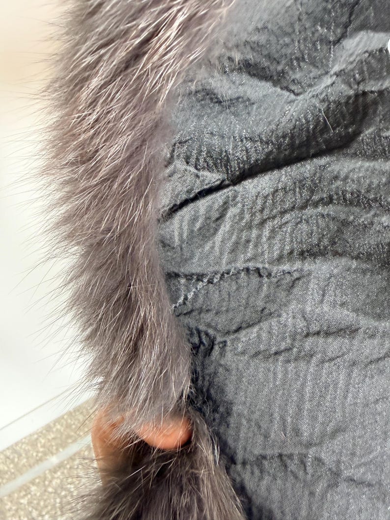 以下が含まれることがあります： テクスチャ加工された表面を持つ濃いグレーの衣服のクローズアップ。ふわふわのライトグレーの毛皮でトリミングされています。毛皮は厚くて柔らかく、生地のしわくちゃの外観とは対照的です。この衣服はアウターウェアの一部のようです。