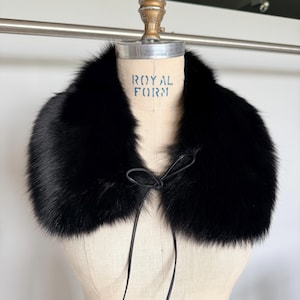 May include: A luxurious black fur collar with a leather tie closure. The collar is displayed on a vintage dress form with the text "ROYAL FORM" printed on the front. The fur appears soft and plush.