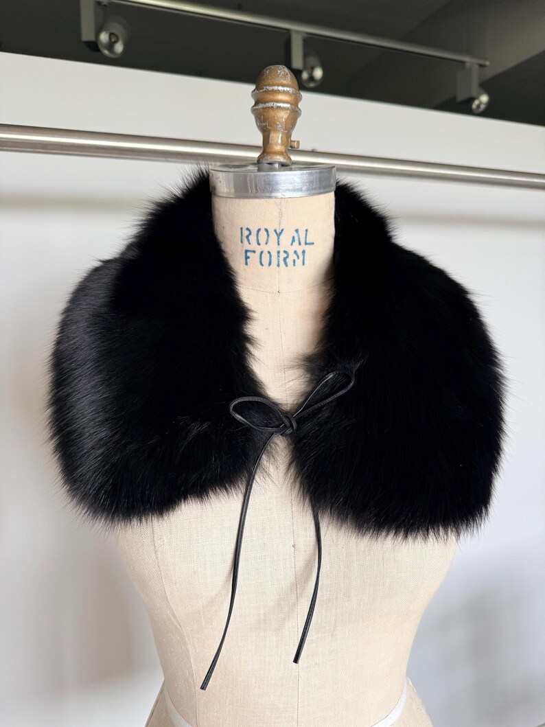 May include: A luxurious black fur collar with a leather tie closure. The collar is displayed on a vintage dress form with the text "ROYAL FORM" printed on the front. The fur appears soft and plush.