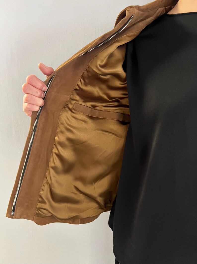 May include: A brown suede jacket with a zip fastening. The interior lining is a golden-brown satin. The jacket has a classic design and a tailored fit, suitable for various occasions.