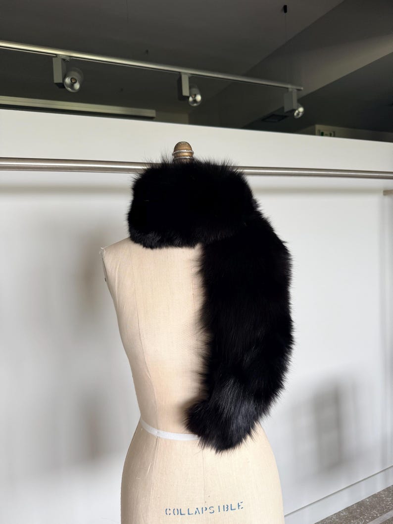May include: A luxurious black fur scarf draped over a mannequin. The scarf is made of soft, dense fur, and it is a deep, solid black color. The mannequin is a light beige color, and the background is a plain white wall.