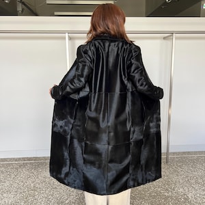 May include: A long, black coat with a glossy finish. The coat is open, revealing the interior lining. The coat's design features a patchwork pattern. The person is wearing beige pants.