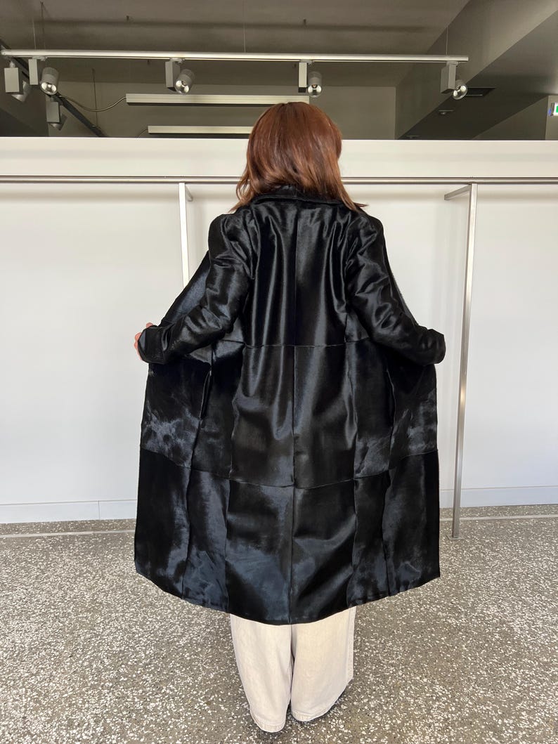 May include: A long, black coat with a glossy finish. The coat is open, revealing the interior lining. The coat's design features a patchwork pattern. The person is wearing beige pants.