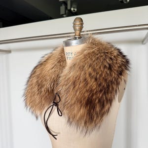 Op de afbeelding: Een luxe bontkraag in bruin- en beigetinten, weergegeven op een mannequin. De kraag heeft een zachte, donzige textuur en is vastgemaakt met een donkerbruin strikje. Een vintage modeaccessoire.