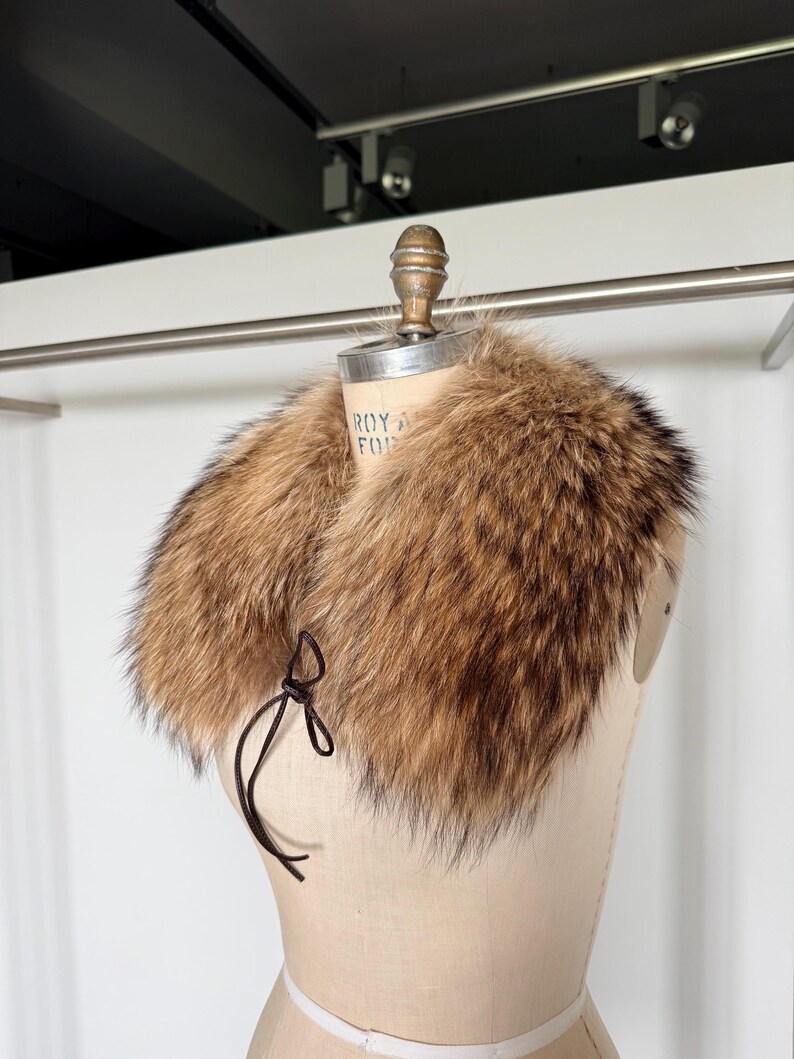 Op de afbeelding: Een luxe bontkraag in bruin- en beigetinten, weergegeven op een mannequin. De kraag heeft een zachte, donzige textuur en is vastgemaakt met een donkerbruin strikje. Een vintage modeaccessoire.