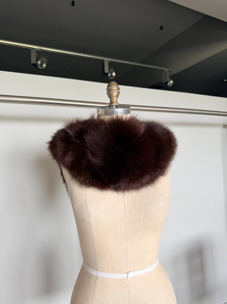 May include: A dark brown fur collar displayed on a mannequin. The collar is a rich, deep shade of brown, with a fluffy texture. The mannequin is a neutral beige color, and the background is a plain white.