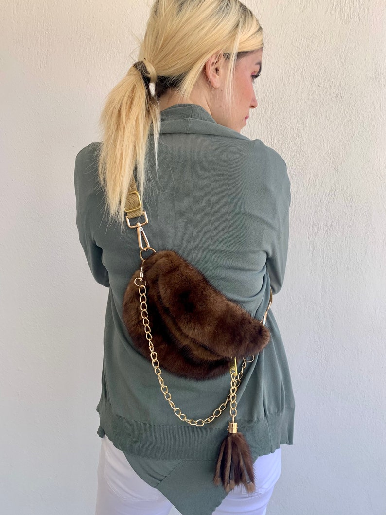 May include: A brown faux fur fanny pack with a gold chain strap and a brown fur tassel. The bag is worn by a person wearing a green sweater and white pants.