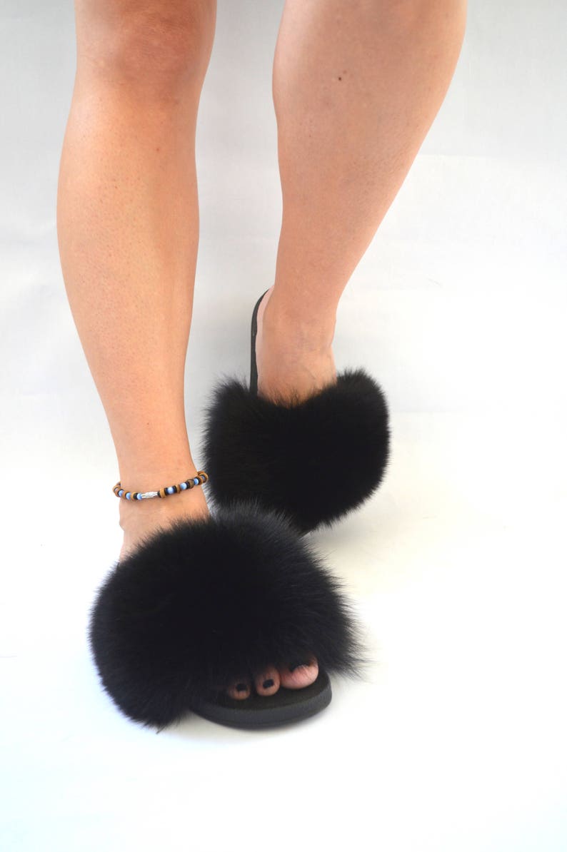 K&ouml;nnte beinhalten: Schwarze Pelz-Sandalen mit flauschigem, dickem Pelz-Futter. Die Sandalen haben einen einfachen schwarzen Riemen und eine schwarze Sohle.