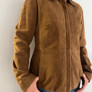 May include: A brown suede jacket with a collared neckline and long sleeves. The jacket has a button-up front and a tailored fit. The model is wearing blue jeans. The jacket is a warm, neutral colour.