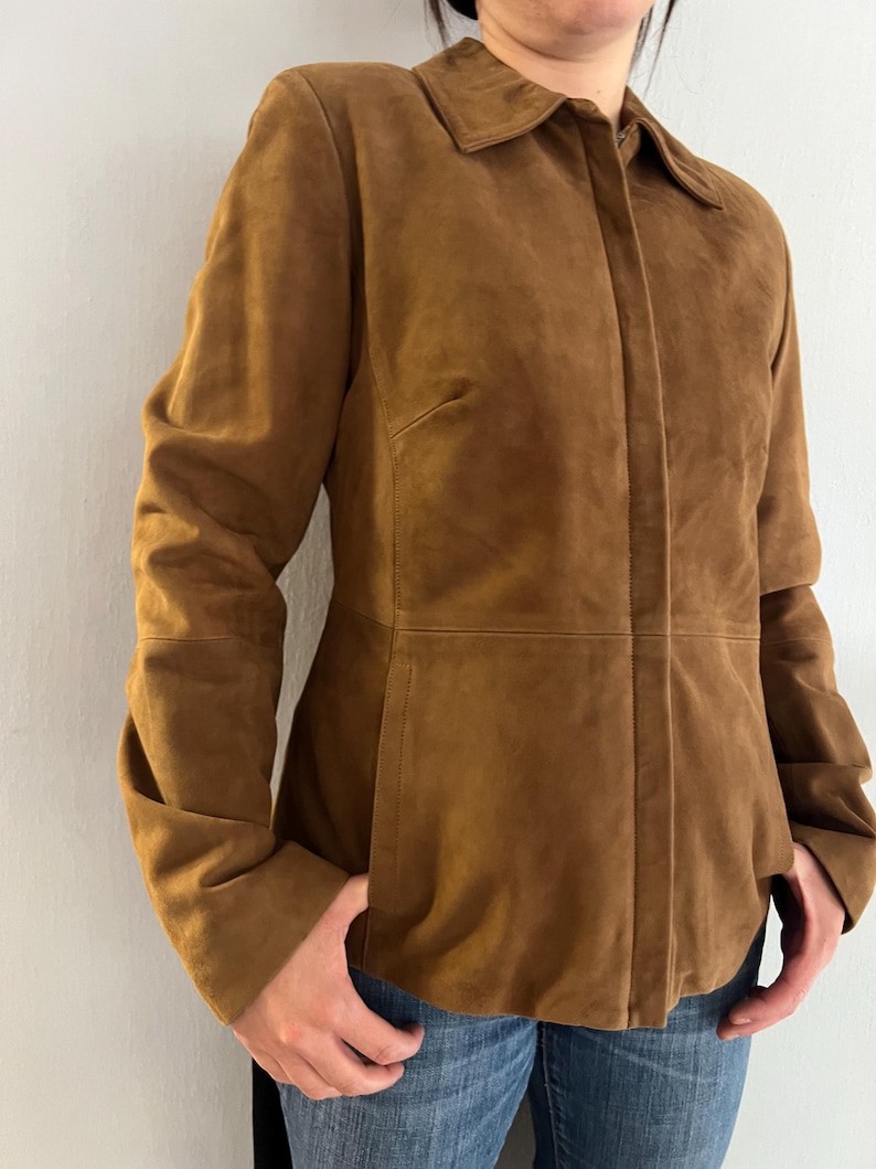 May include: A brown suede jacket with a collared neckline and long sleeves. The jacket has a button-up front and a tailored fit. The model is wearing blue jeans. The jacket is a warm, neutral colour.