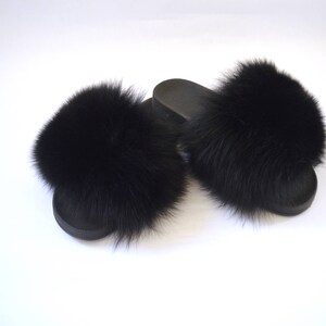 K&ouml;nnte beinhalten: Ein Paar schwarze Pelz-Sandalen. Die Sandalen haben an jedem Riemen einen flauschigen schwarzen Pelz-Bommel.