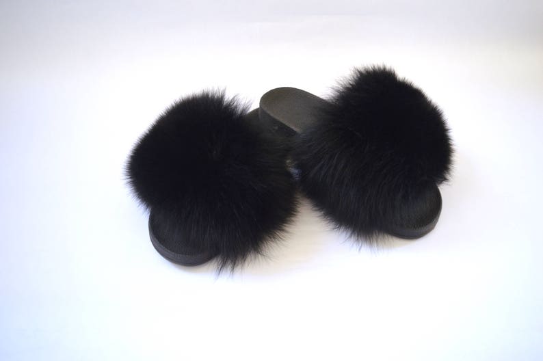 K&ouml;nnte beinhalten: Ein Paar schwarze Pelz-Sandalen. Die Sandalen haben an jedem Riemen einen flauschigen schwarzen Pelz-Bommel.