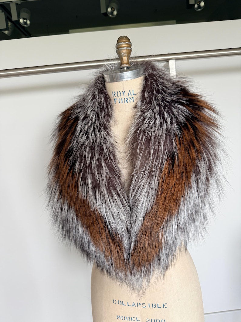 May include: A luxurious fur collar with a mix of brown, silver, and orange tones. The collar is displayed on a mannequin, showcasing its shape and texture. The fur appears soft and fluffy, perfect for adding a touch of elegance to any outfit.