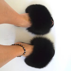 K&ouml;nnte beinhalten: Ein Paar schwarze Fellsandalen mit einem flauschigen Bommel-Detail. Die Sandalen werden an den F&uuml;&szlig;en einer Person getragen, und ein Perlen-Fu&szlig;kettchen ist sichtbar.