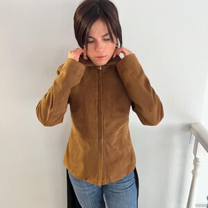 May include: A brown suede jacket with a zip fastening. The jacket has a fitted shape and long sleeves. The person in the image is wearing blue jeans.
