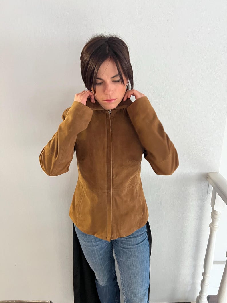 May include: A brown suede jacket with a zip fastening. The jacket has a fitted shape and long sleeves. The person in the image is wearing blue jeans.