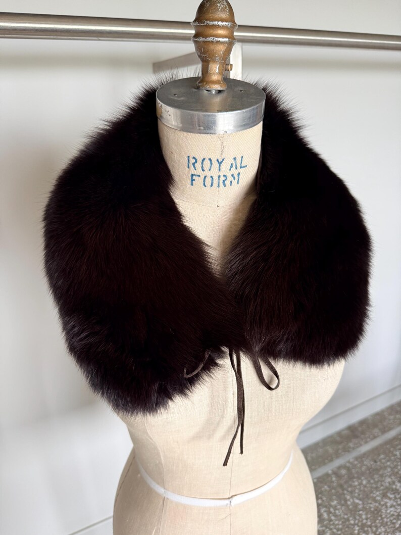 May include: A dark brown fur collar with a tie closure, displayed on a vintage dress form. The collar is plush and appears to be made of luxurious fur. The dress form has the text "ROYAL FORM" printed on it.