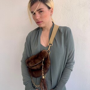 May include: A woman wearing a sage green cardigan with a brown fur fanny pack with a gold chain and tassel. The fanny pack is worn across the body.
