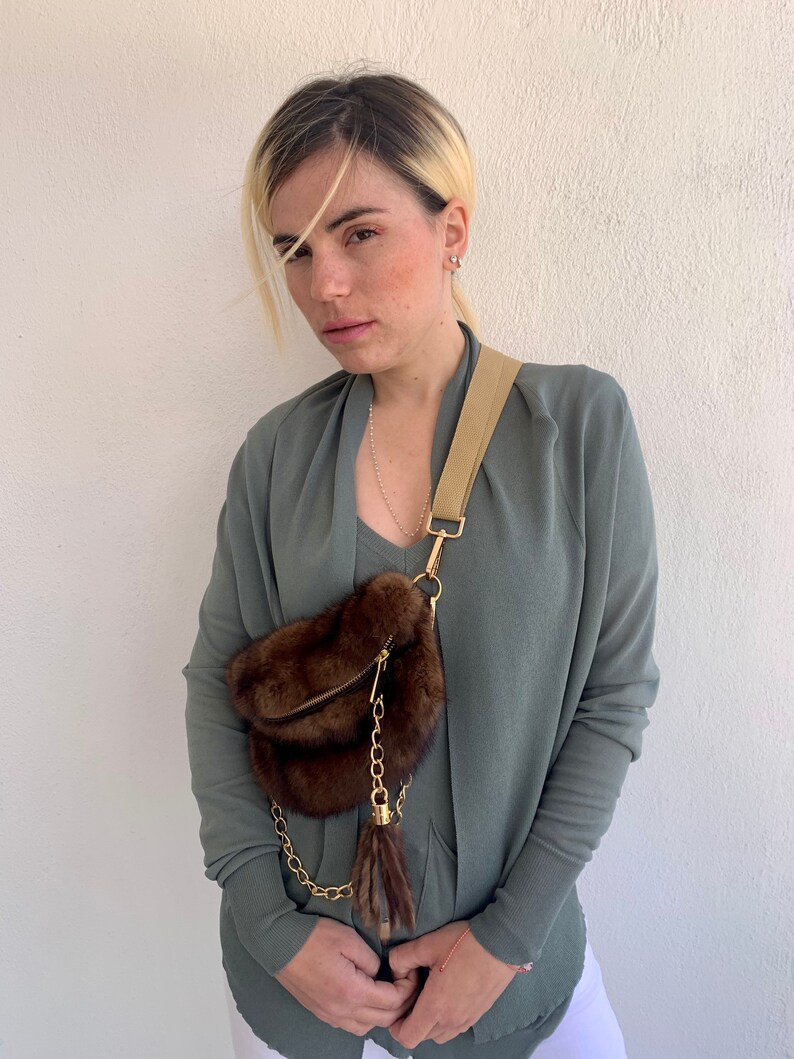 May include: A woman wearing a sage green cardigan with a brown fur fanny pack with a gold chain and tassel. The fanny pack is worn across the body.