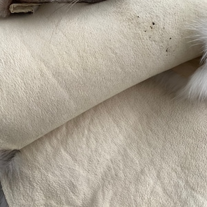 Puede incluir: Primer plano de una tela de piel beige claro con una superficie ligeramente texturizada. La tela est&aacute; doblada por la mitad, revelando la parte inferior, que es de color beige claro liso.