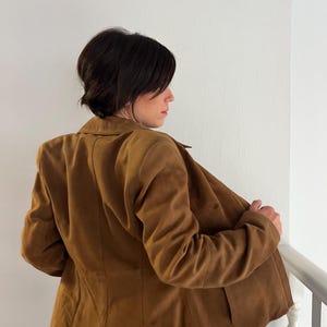 May include: A brown suede blazer with a classic collar and tailored fit. The jacket has a button closure and two front pockets. The model is wearing the blazer, with the back facing the camera, and is holding the lapel open.