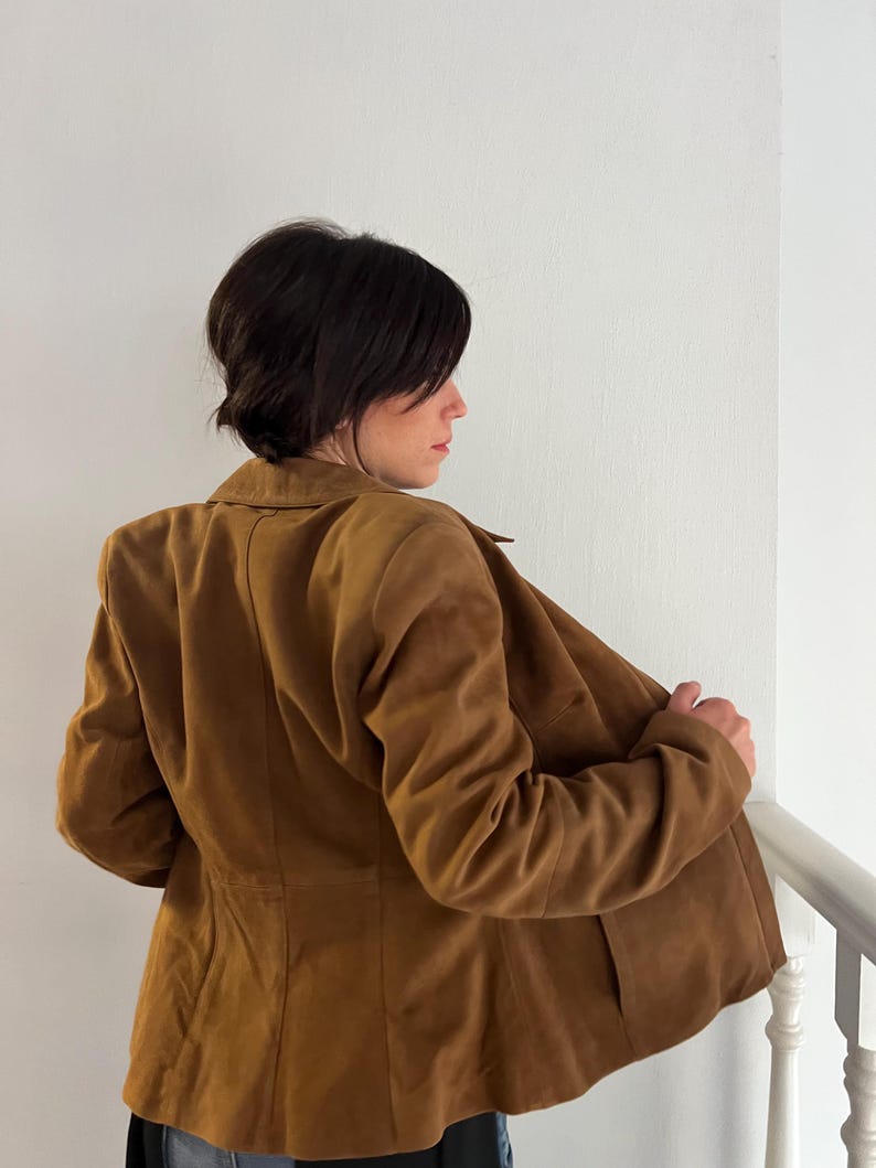 May include: A brown suede blazer with a classic collar and tailored fit. The jacket has a button closure and two front pockets. The model is wearing the blazer, with the back facing the camera, and is holding the lapel open.