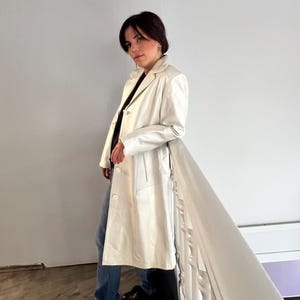 Könnte beinhalten: Ein cremefarbener, langer Mantel mit Knopfleiste vorne. Der Mantel hat einen klassischen Reverskragen und eine taillierte Passform. Die Person trägt blaue Jeans und schwarze Schuhe. Der Hintergrund ist eine weiße Wand und eine weiße Treppe.