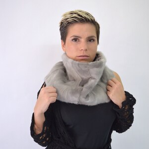 May include: A woman with short blonde hair is wearing a grey fur scarf and a black crocheted cardigan.