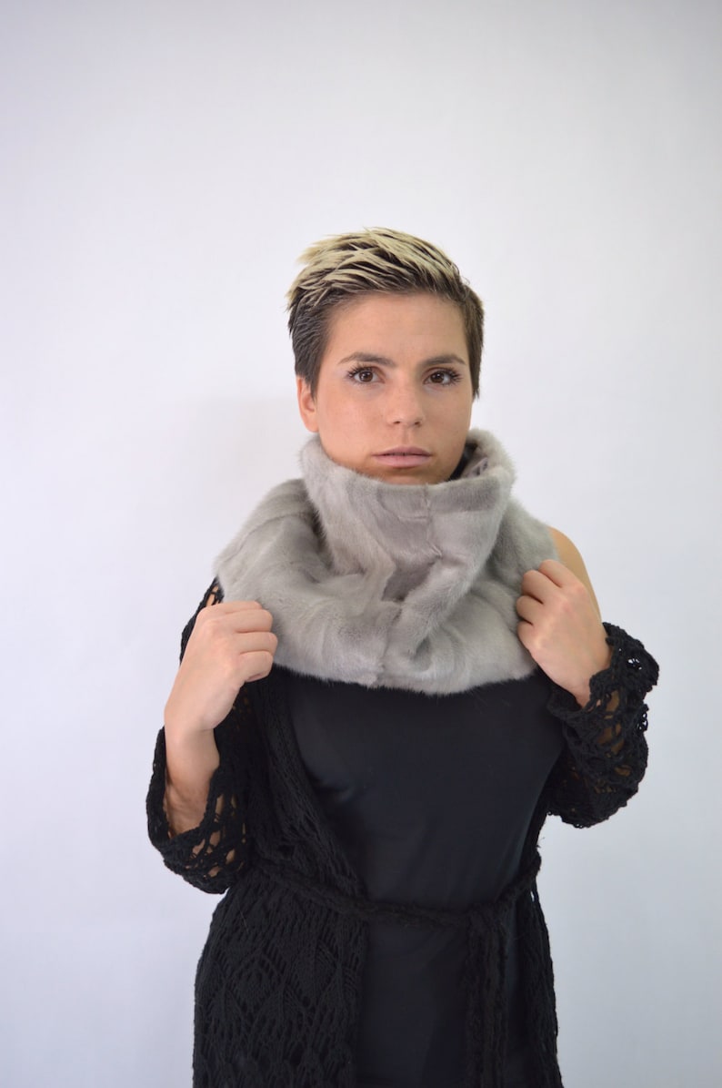May include: A woman with short blonde hair is wearing a grey fur scarf and a black crocheted cardigan.