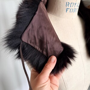 May include: A vintage fur collar with a dark brown satin lining and a black fur trim. The collar is held by a hand, showcasing the texture and color of the fur. A brown leather tie is attached to the collar.