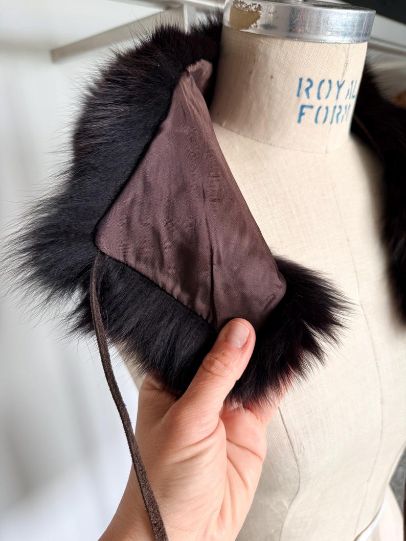 May include: A vintage fur collar with a dark brown satin lining and a black fur trim. The collar is held by a hand, showcasing the texture and color of the fur. A brown leather tie is attached to the collar.